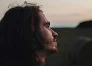 photo-realistic image of a medium-light-complexioned young adult man in profile. The man has long, wavy brown hair, and his gaze appears contemplative. He is outside, and behind him is a light, twilight sky.